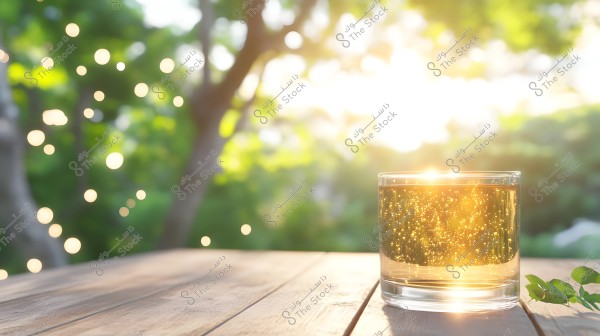 The image shows a clear glass containing a fizzy yellow beverage, placed on a wooden table outdoors. The sunlight from the background fills the image with warm light, reflecting golden bubbles from the drink. In the blurred background, there is a vague view of green foliage and circular bokeh lights.