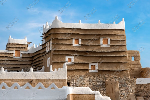 A traditional mud-and-brick building with a unique architectural design located in Saudi Arabia. The brown walls have horizontally protruding layers and feature rectangular wooden windows. The roof is coated in white, adding aesthetic appeal and decorative charm to the structure. The clear blue sky enhances the visual details of the traditional building.
