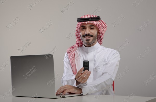 Image of a man sitting at a desk wearing traditional Saudi attire, consisting of a white thobe and a red and white checkered shemagh with a black agal. He appears happy while working on a laptop and holding a remote control in his right hand.
