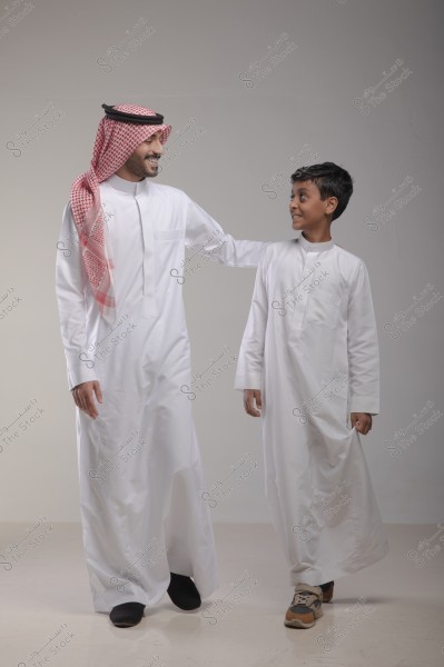 A man and a boy stand smiling in a photography studio. The man is wearing a white thobe and a red-checkered shemagh, while the boy is wearing a white thobe and sneakers. The man gently places his hand on the boy\'s shoulder. The background is simple and neutral in color.