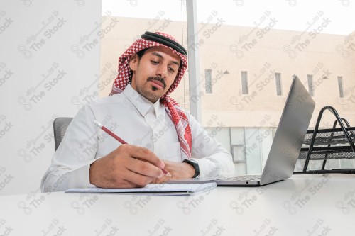 رجل سعودي يرتدي الزي السعودي التقليدي يعمل في مكتب بواجهة زجاجية ويستخدم قلم رصاص لتدوين الملاحظات