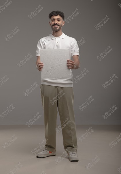 Image of a man standing against a grey background, wearing a white polo shirt and beige trousers with sneakers. The man is holding a rectangular white board with both hands. It appears as a studio or promotional shot.