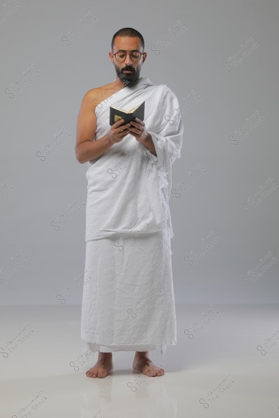 A portrait of a man standing in a contemplative pose, wearing a traditional white Ihram, worn for Hajj and Umrah, and reading from a small book. The background is gray, suggesting simplicity and calmness.