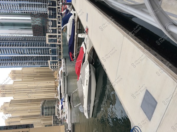 View of Dubai Marina, featuring a collection of luxury yachts moored beside a wooden dock. The modern cityscape is prominent in the background with impressive high-rise skyscrapers. The yachts are covered with vibrant-colored covers, reflecting in the calm water.