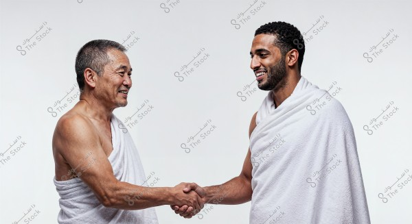 The image shows two men shaking hands with smiles. Both are wearing white Ihram clothing, and the background is white and neutral.