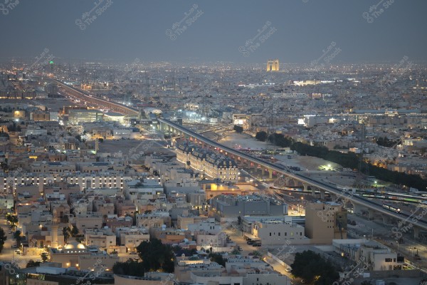 Aerial view of Riyadh at night, showing a collection of residential and commercial buildings illuminated with artificial lights. A highway is visible running through the image, with a few cars on it. In the background, the city lights extend to the horizon.