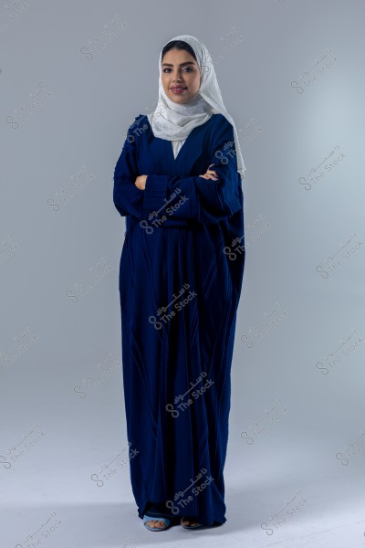 A portrait of a woman wearing a blue abaya and a white headscarf, standing with her arms crossed in front of her. The background is gray, and the woman is looking forward with a slight smile.