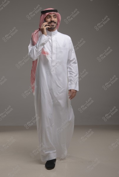 A portrait of a man wearing a traditional white thobe with a red-checkered shemagh and agal on his head, talking on a mobile phone. He stands against a neutral gray background. The attire reflects traditional Saudi clothing.