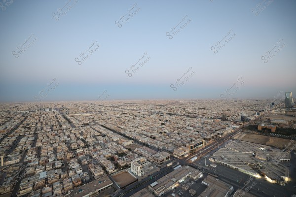 Aerial view of a large city in Saudi Arabia, showing a grid of houses and buildings under a clear blue sky at sunset. A tall tower is visible on the right side of the image, likely the Kingdom Tower in Riyadh. The buildings are arranged in a regular pattern with wide roads separating them.