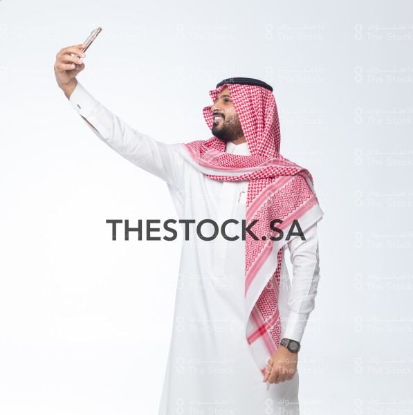 بورتريه رجل سعودي على خلفية بيضاء يقوم بالتقاط صور سيلفي وهو مبتسم ، صور تذكارية ، توثيق لحظة سعيدة