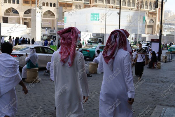 The image shows two men wearing traditional white robes and red ghutras walking down a busy street near the Grand Mosque in Mecca. In the background, several people dressed in traditional white attire are sitting near cars and buildings with traditional architectural designs.