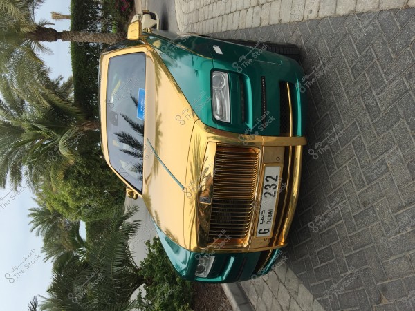 The image shows a luxury car in metallic gold and green colors parked next to a curb in an area lined with palm trees. The car has an Emirati license plate with symbols indicating Dubai. The weather appears to be sunny, highlighting the shine of the gold coating on the car.