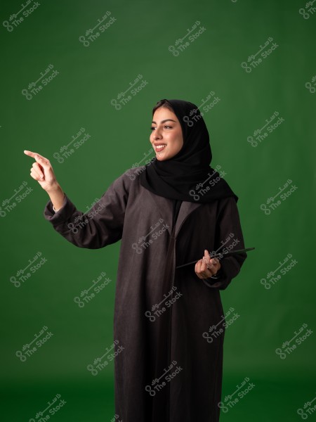 An image of a woman wearing a black abaya and black hijab, pointing to the right with her right hand while holding a tablet in her left hand. The background is green.
