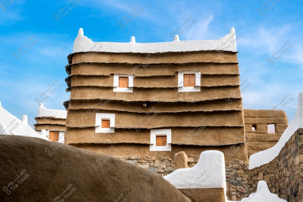 A traditional mud-and-brick building with a unique architectural design located in Saudi Arabia. The brown walls have horizontally protruding layers and feature rectangular wooden windows. The roof is coated in white, adding aesthetic appeal and decorative charm to the structure. The clear blue sky enhances the visual details of the traditional building.