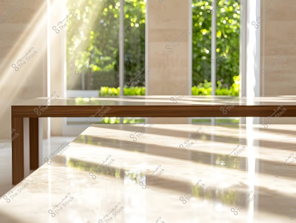 An interior view of a room illuminated by sunlight streaming through large windows. The image shows a modern table made of wood and glass, reflecting the natural light. Behind the window, there is a view of a lush green garden filled with leafy trees.