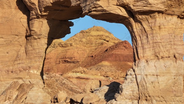 A natural landscape of a rock formation forming a natural arch in the desert with a mountain in the background under a blue sky.