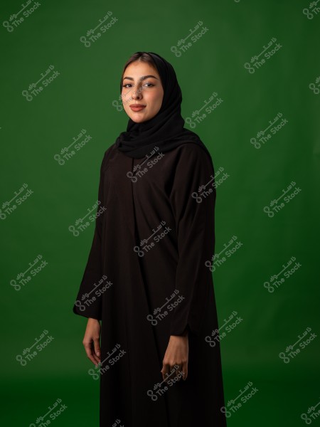 امرأة ترتدي حجاباً وعباءة سوداء تقف أمام خلفية خضراء.