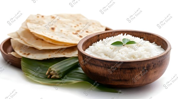 An image of a wooden bowl filled with white rice, topped with two green leaves. Next to the bowl, there\'s a stack of flatbread placed on green leaves.