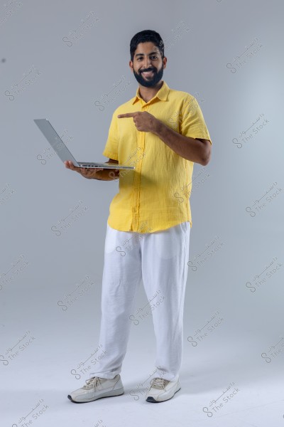 A portrait of a smiling man standing, wearing a yellow shirt, white pants, and white sneakers, holding a laptop in one hand and pointing at it with the other. The background is simple with a light gray color.