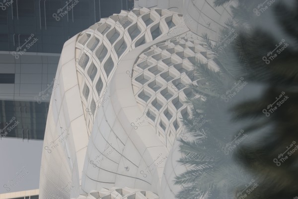 صورة لقسم من مبنى حديث بتصميم معقد, يتميز بزخارف مثلثية متشابكة وواجهات منحنية. في المقدمة, تظهر أشجار نخيل غامضة ويظهر في الخلفية جزء من مبنى زجاجي.