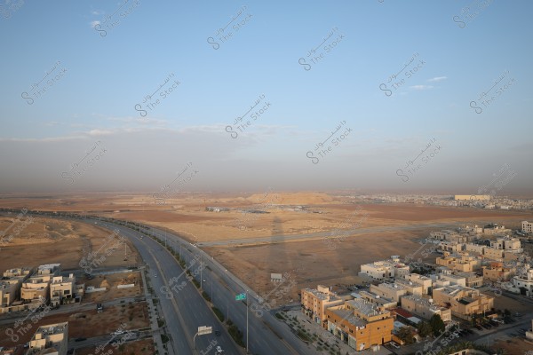 The image shows an aerial view of a desert landscape with residential buildings. A main road runs through the picture with a few cars, and modern houses are scattered to the right. The sky is clear with a few scattered clouds.