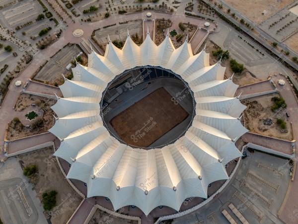 An aerial view showing a circular architectural design of a building resembling a tent with a polygonal roof, situated in an open area with surrounding parking lots and scattered trees. The structure consists of a series of uniform white conical shapes forming a large circle.