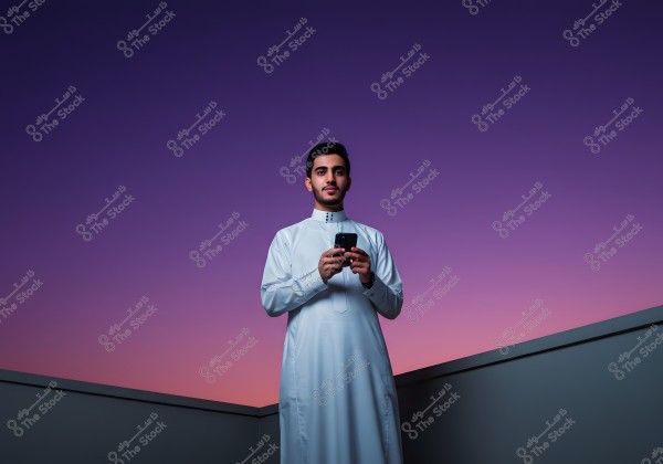 A portrait of a man standing on a rooftop at sunset. The background features a sky with gradients of purple and pink. The man is wearing traditional white attire and is using a mobile phone.