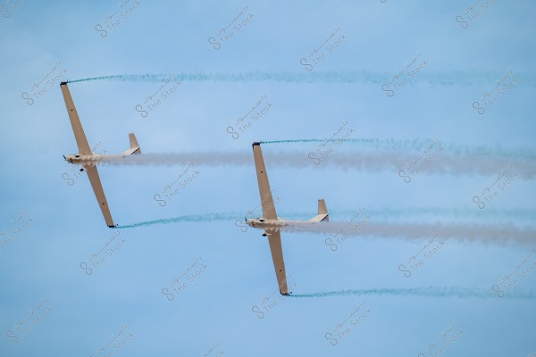 Two glider planes flying in the blue sky, leaving colored smoke trails behind them. The plane on the left is slightly tilted upwards, and the plane on the right is slightly tilted downwards. The smoke trails are white and light blue.