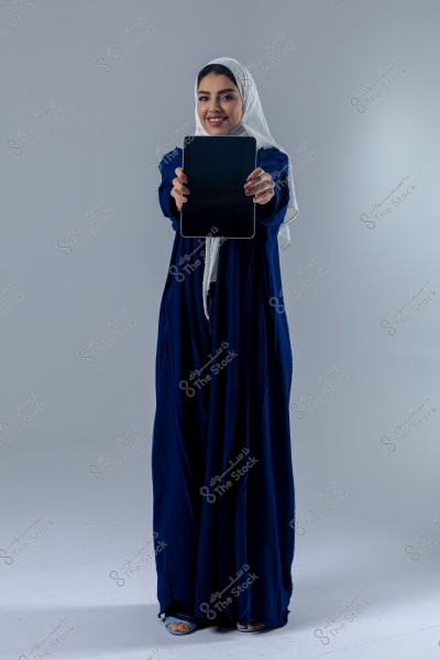 A portrait of a woman wearing a blue abaya and a white hijab, holding a tablet in her hands in front of her against a gray background. The image suggests she is from the Middle East, possibly Saudi Arabia. The overall design is simple, with an elegant and modest appearance.