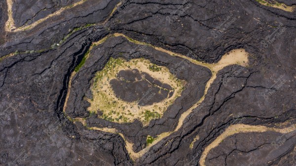 An aerial view of the earth\'s geology showing black rock formations in ring-like shapes surrounded by sandy terrain. The land\'s topography is clearly visible with color gradients ranging from black to light brown, with small green patches representing desert vegetation.