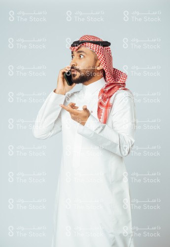 بورتريه رجل سعودي يرتدي الزي السعودي التقليدي يتحدث بالجوال بينما يشير بيده وهو يتحدث، شاب مندمج بمكالمة هاتفية