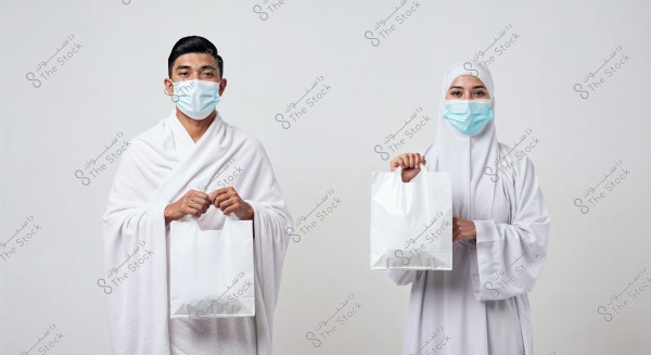 The image shows a man and a woman wearing white Ihram clothing, wearing blue surgical masks, each holding a white paper bag. The attire suggests that the scene might be related to Hajj or Umrah. They stand against a plain white background.