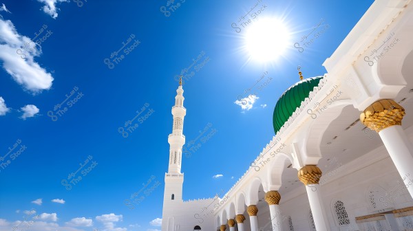 An image of the Prophet\'s Mosque in Medina, Saudi Arabia. The picture shows the distinctive green dome and a tall white minaret under the clear blue sky with a bright sun. Part of the mosque\'s structure is visible, featuring white and gold columns.