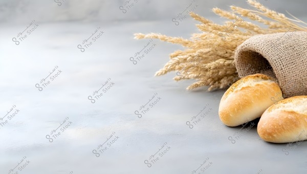 The image shows two round loaves of bread on a light gray surface next to a burlap sack containing stalks of golden wheat. The natural arrangement evokes a rustic and calm atmosphere.