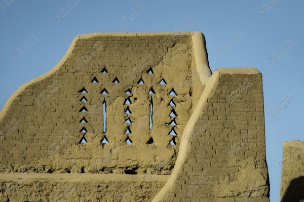Image of an old mud wall reflecting traditional architecture. The wall features triangular-shaped openings arranged in rows, with a distinctive curve in the design at the upper left side. The clear blue sky forms a backdrop for the wall.