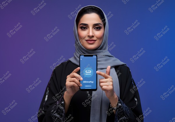 A woman wearing an embellished black abaya and a gray scarf, standing against a gradient blue to purple background. She is holding a smartphone displaying the \"AlinnaPay\" app and pointing towards the phone\'s screen with her other hand.