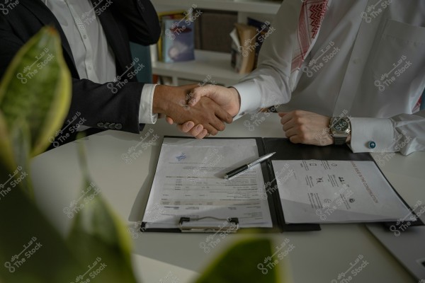 The image shows two individuals shaking hands over a desk. The person on the right is wearing a white thobe and a red-checkered shemagh, with a watch on the wrist, suggesting they might be from the Gulf region. The other person is wearing a black suit and a white shirt. There are several papers on the desk in front of them, along with a pen. In the background, a bookshelf and a green plant are visible.