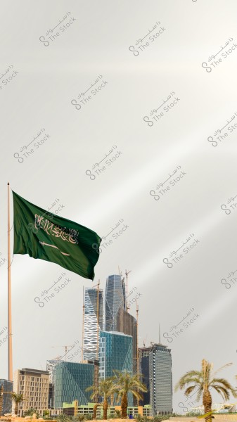تظهر الصورة علم المملكة العربية السعودية الأخضر يرفرف في الهواء. في الخلفية, توجد مجموعة من المباني الحديثة وناطحات السحاب تحت الإنشاء, محاطة بأشجار النخيل. السماء خلف العلم تبدو صافية.