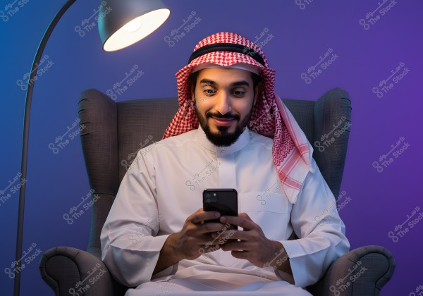 A photo of a man sitting on a comfortable chair under a floor lamp. He is wearing traditional Saudi attire with a white thobe, red and white ghutra, and agal. The man is using a phone and looks pleased. The background is lit with purple and blue hues, contrasting with the soft lighting of the lamp.