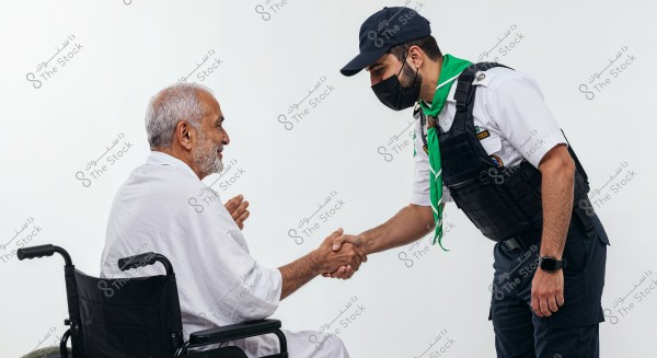 The image shows an elderly man sitting in a wheelchair wearing a white Ihram, shaking hands with another person dressed in a scouting uniform with badges. The scout is wearing a white shirt, blue trousers, a tactical vest, a green scarf, and a cap. The background is completely white, emphasizing the interaction between the two individuals.