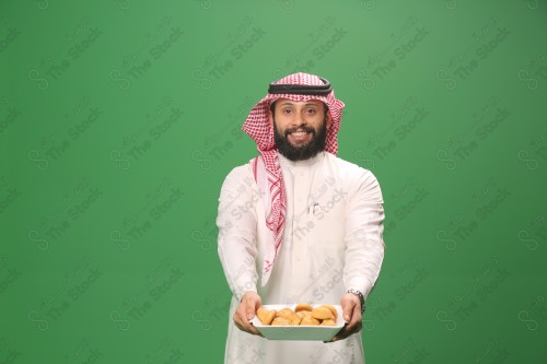 شاب سعودي على خلفية خضراء يرتدي الثوب و الشماغ، يقوم بعمل حركات بيده،يمسك بصينية الكعك و يقدمه، احتفالات العيد