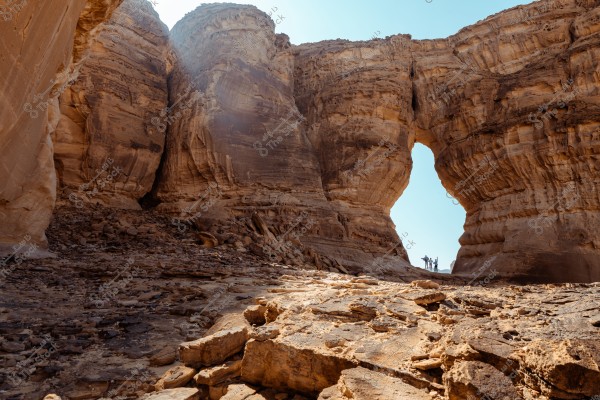 A natural landscape showing massive and tall rock formations in a desert area. The rocks exhibit various shades of brown extending towards the blue sky. In the center of the rock formations, there is an arch-shaped opening where three people are standing at the edge, appearing small compared to the massive size of the rocks.