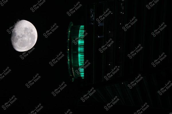 An image showing a bright full moon on the left, and on the right, part of a building illuminated with green lights. The visible section of the building has a modern design and contrasts with the dark night sky.