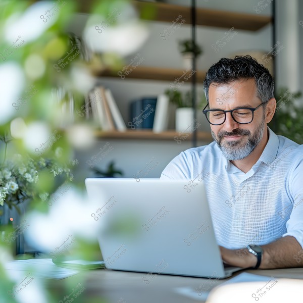 An image of a man sitting in a home office, working on a laptop. The man is wearing a light blue shirt and glasses. In the background, there are shelves with books and decorative plants. The setting appears relaxed, with the man working in a bright, naturally lit workspace, and plants in the foreground.
