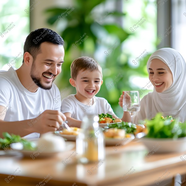 A family of three gathers around a dining table in a well-lit home. The father, mother, and young child are wearing casual white clothing and enjoying a healthy meal that includes vegetables. The mother is wearing a white hijab, suggesting an Eastern background. The atmosphere is cozy and cheerful, with green plants in the background adding a sense of warmth and relaxation.