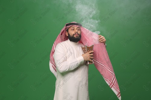 رجل سعودي يقف على خلفية خضراء يرتدي الثوب و الشماغ، يقوم بعمل حركات بيديه ، يبخر نفسه بالطيب و العود، احتفالات العيد