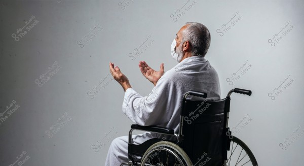 An elderly man sitting sideways in a wheelchair, wearing a white robe and a face mask, raising his hands in prayer. The background is plain and white.