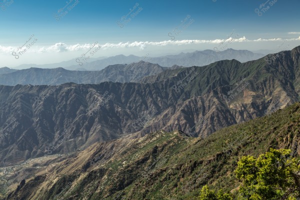 Aerial view of a mountain range under a clear blue sky with some white clouds on the horizon. The mountains are covered with green and brown, creating a stunning natural impression.