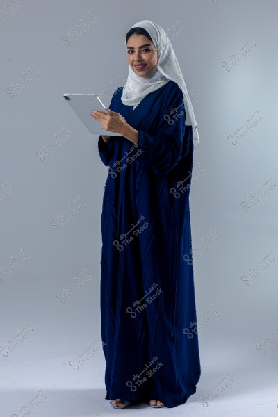 A portrait of a woman wearing a blue abaya and a white hijab, standing and smiling while holding a tablet. The outfit suggests it may be of Saudi style.