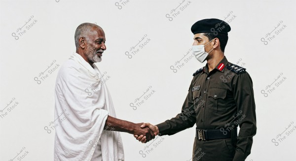 An image of two men shaking hands. The man on the left appears elderly and is wearing a white Ihram, traditional attire for the Hajj and Umrah rituals. The man on the right is a police officer in a dark-colored uniform, wearing a cap and a face mask. Both stand in front of a white background.